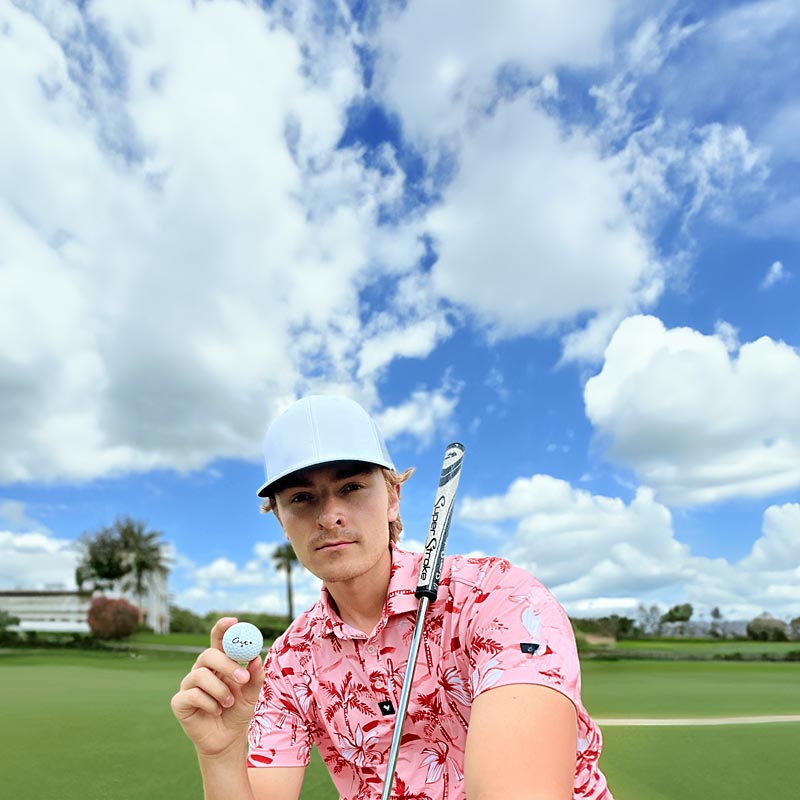 Golfer With a White Hat On Holds a Cut Golf Ball As He Takes a Selfie